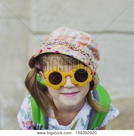 Cute smiling happy little girl with a backpack sitting on a bench. Child tourist in big old city. Kid in sunglasses during summer vacation. Happy little traveler with ponytails. Copy space.