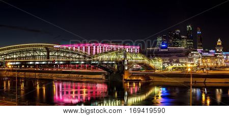 Night winter panorama of the Moskva River embankment: pedestrian bridge Bogdan Khmelnitsky Kiyevskaya railway station Moscow-City Redisson Slavyanskaya hotel. Rich colorful illuminations and reflections in the river.