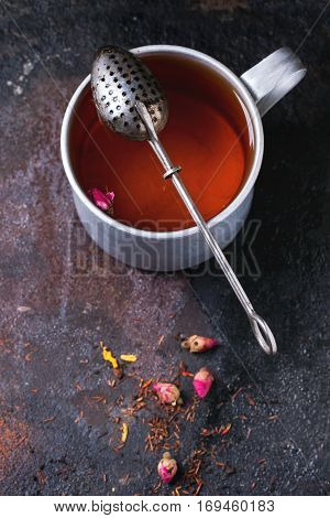 Vintage aluminum mug of hot tea with tea strainer, dry tea and rose buds over dark background. Top view