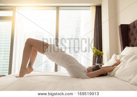 Young sporty attractive woman practicing yoga at home or hotel bedroom after waking up, stretching in dvi pada pithasana exercise, Glute Bridge, working out wearing white home wear, indoor full length