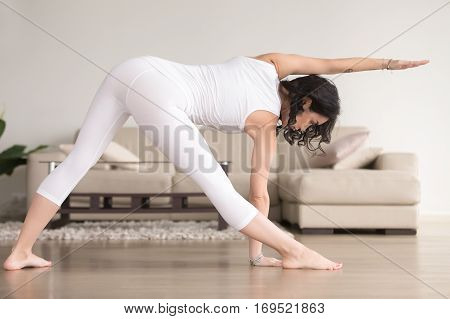 Young attractive woman practicing yoga at home, standing in extended triangle exercise, Utthita Trikonasana pose, working out, wearing white clothes, indoor full length, living room. Back view