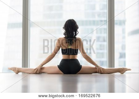 Young sporty woman practicing yoga, sitting in Samakonasana exercise, Straight Angle pose, working out, wearing sportswear, black tank top, shorts, indoor full length, against floor window, rear view