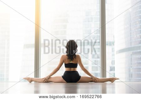 Young sporty woman practicing yoga, sitting in Samakonasana exercise, Straight Angle pose, working out wearing sportswear black tank top, shorts, indoor full length, rear view. Copy space
