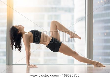 Young attractive woman practicing yoga, standing in Upward Plank exercise, Purvottanasana pose, working out, wearing sportswear, black tank top, shorts, indoor full length, floor window with city view
