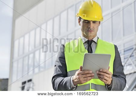 Young male engineer in reflector-vest and hardhat using digital tablet outside industry
