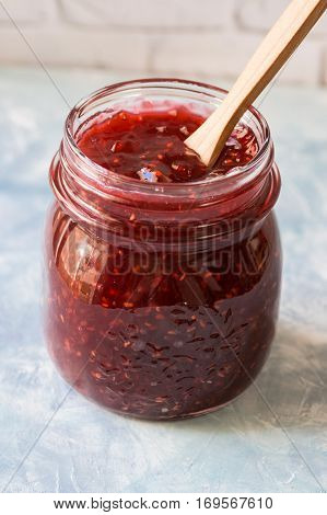 Jar with homemade raspberry jam, with dipped wooden spoon, on concrete stone table, brick wall in the background, kitchen interior, making of preserves, breakfast