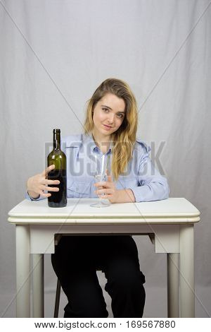 Young Caucasian white woman sitting at a table with a glass of wine in her hand and on the other a bottle of wine. She is blond and pretty with light brown eyes. The background of the image is white.