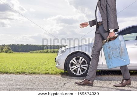 Low section of young businessman with gas can walking by broken down car at countryside