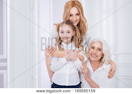 Family life. cheerufl content loving woman embracing her little daughter and snior mother while resting together at home and spending time with pleasure