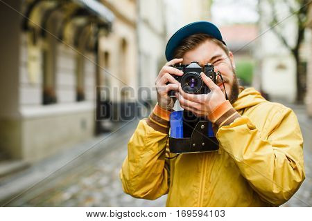 Young hipster with a beard holding oldshool film camera and ready to take a photo, on the street, wearing in bright raincoat and hat, portrait, day outdoors, closeup