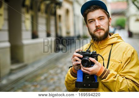 Young hipster with a beard holding oldshool film camera, thinking and ready to take a photo, on the street, wearing in bright raincoat and hat, portrait, day outdoors, closeup, soft-focus