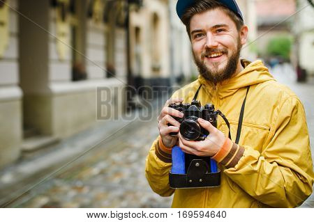Young hipster with a beard holding oldshool film camera, laughing and ready to take a photo, on the street, wearing in bright raincoat and hat, portrait, day outdoors, closeup