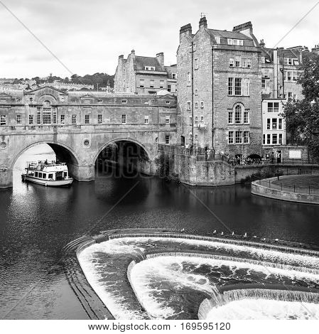 Bath, Somerset, United Kingdom - June 19, 2006: A river cruise tour boat ferries passengers along the River Avon. The Pulteney Bridge (Georgian architecture) over the river Avon with weir in Bath. Black and white.