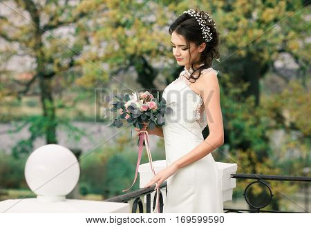Closeup portrait of young gorgeous bride. Pretty beautiful bride with beautiful bouquet of flowers standing in balcony of amazing mansion on spring tress background. Pretty bride with hairstyle.