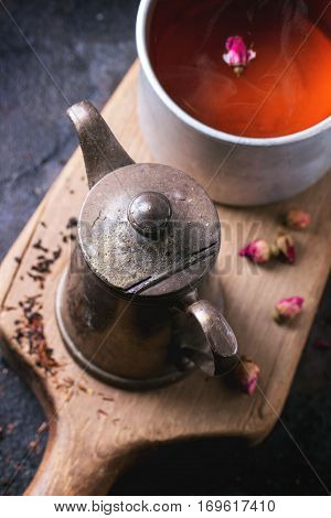 Vintage teapot and aluminum mug of tea, served on wooden cutting board with dry rose buds over dark background. Top view