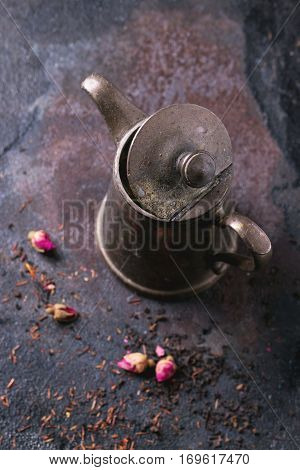 Open vintage teapot with dry tea and rose buds over black metal background. Top view