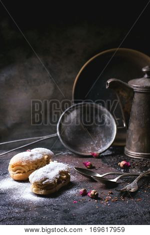Tea drinking with eclairs and chopped chocolate, served with vintage teapot and cutlery over dark background. See series