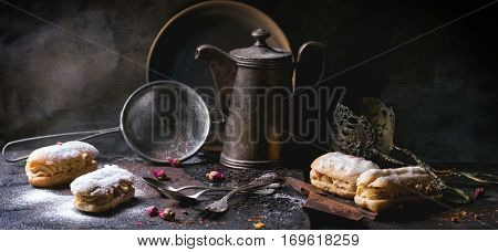 Tea drinking with eclairs and chopped chocolate, served with vintage teapot and cutlery over dark background. See series