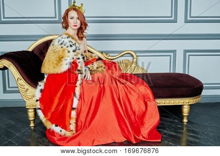 Red-haired woman in red dress, cloak and with crown on head sits on couch.