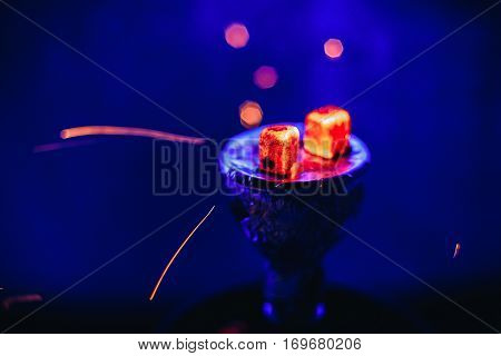 Shisha hookah with glowing red embers and flying sparks in the bowl on blue background