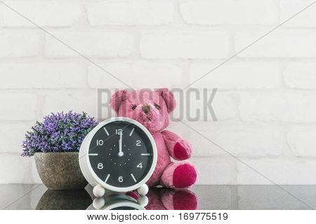 Closeup black and white alarm clock for decorate in 12 o'clock with bear doll and plant on black glass table and white brick wall textured background with copy space