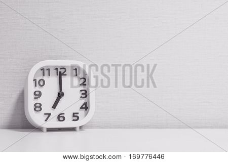 Closeup white clock for decorate in 7 o'clock on white wood desk and cream wallpaper textured background in black and white tone with copy space