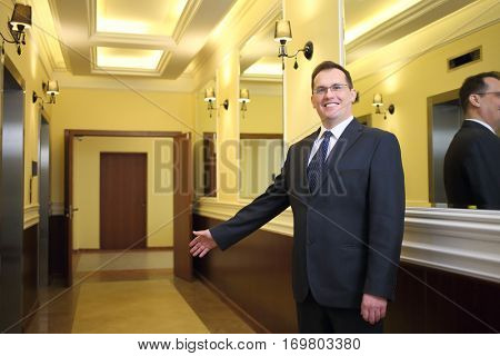 Hospitable concierge in a business suit standing in the hallway between the elevator and mirrors and inviting to enter the house
