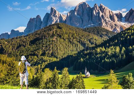 Sunny day in Dolomites, Tirol. Active elderly woman-tourist with backpack photographs the church of Santa Maddalena. Forested mountains surrounded by green Alpine meadows