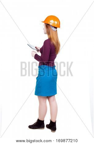 Backview of business woman in construction helmet stands and enjoys tablet or using mobile phone. Isolated over white background. Young woman engineer working on tablet. Engineer scorekeeper at work
