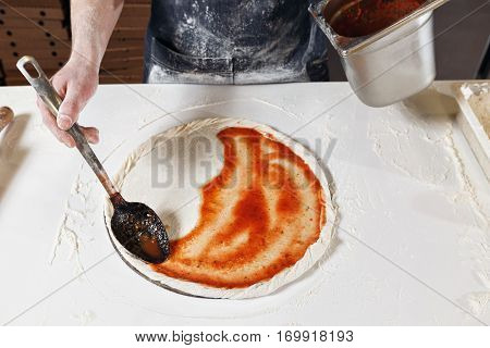 Cooking pizza. the workpiece poured tomato sauce. Closeup hand of chef baker in uniform blue apron cook at kitchen.