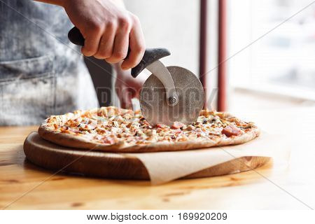 Closeup hand of chef baker in uniform blue apron cutting pizza at kitchen.