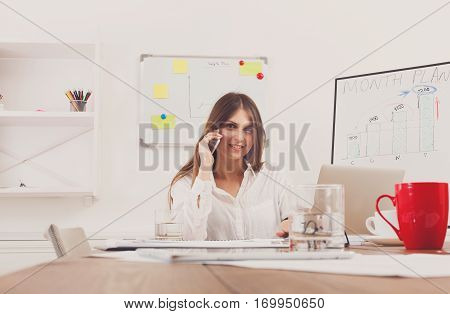 Beautiful young businesswoman talking, answer a call at mobile phone, sitting at wooden desk with laptop and red cup. Modern office worker in white room. Succesfull woman, female carreer concept.