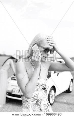 Frustrated woman using cell phone while friend examining broken down car at countryside