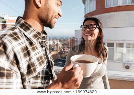 Picture of happy young loving couple drinking coffee while standing on roof. Look at each other.