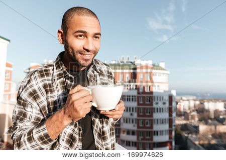 Image of attractive young african man drinking coffee while standing on roof. Look aside.