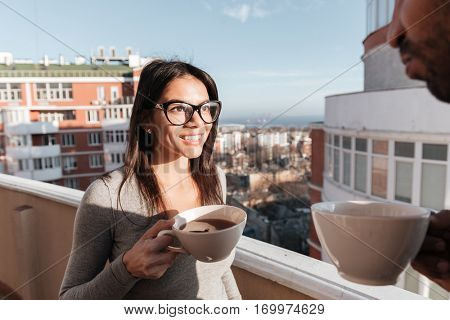 Photo of young loving couple drinking coffee while standing on roof. Look at each other.