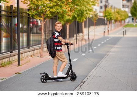 School Boy In Riding With His Electric Scooter In The City With Backpack On Sunny Day. Child In Colo