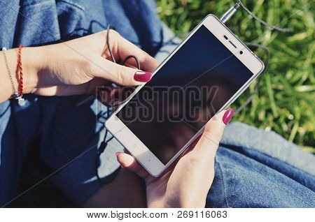 Hands Of A Young Girl Who Holds A Smartphone And Sits On The Green Grass, Close-up