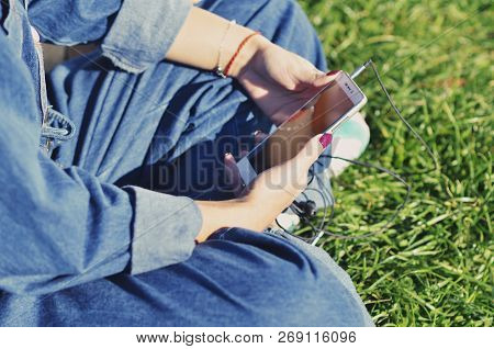Hands Of A Young Girl Who Holds A Smartphone And Sits On The Green Grass, Close-up