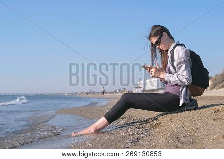 Traveler Girl With A Backpack Sitting On A Sandy Sea Beach And Typing A Message On Mobile Phone.