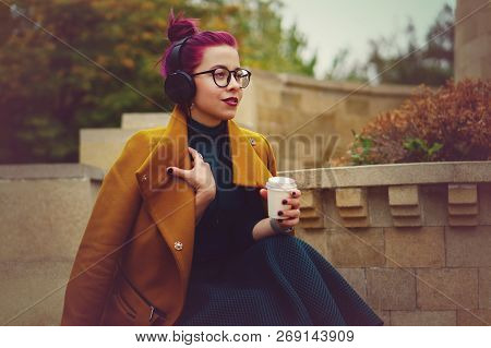 Cute Young Girl Listening To Music On Headphones In Autumn Park. Woman Is Holding Paper Cup With Hot