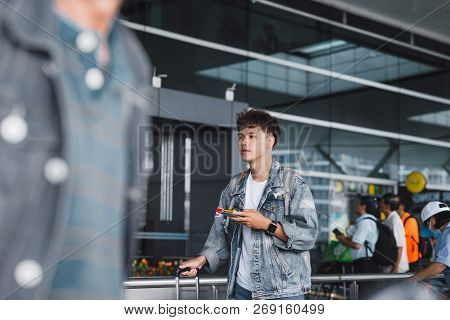 Young Aisan Man Texting On The Phone While Standing Near Rows Of Luggage Trolleys In The Airport