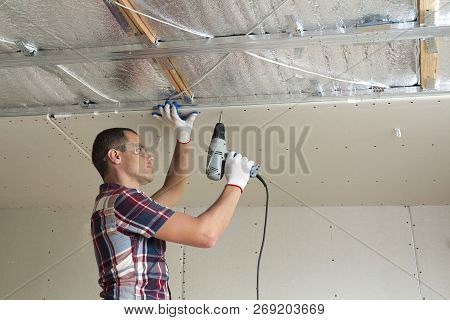 Young Man In Goggles Fixing Drywall Suspended Ceiling To Metal Frame Using Electrical Screwdriver On