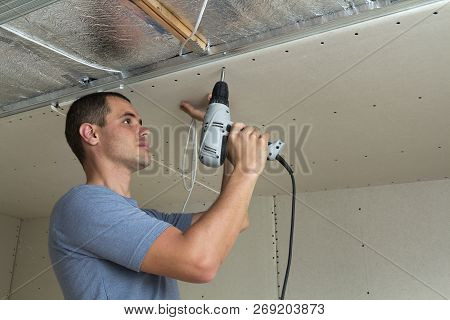 Young Man Uses Electrical Screwdriver Fixing Drywall Suspended Ceiling To Metal Frame On Ceiling Ins
