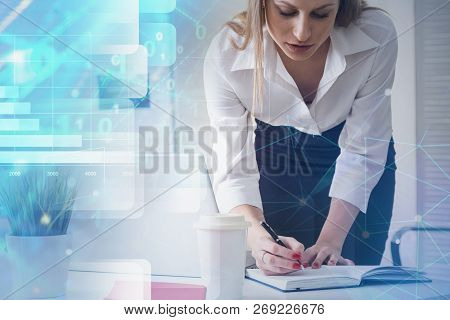 Blonde Businesswoman Writing In Planner In Her Office With Double Exposure Of Digital Interface Holo