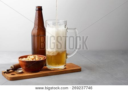 Lager Beer Pouring In Glass And Bottle With Salted Peanuts On Gray Concrete Background