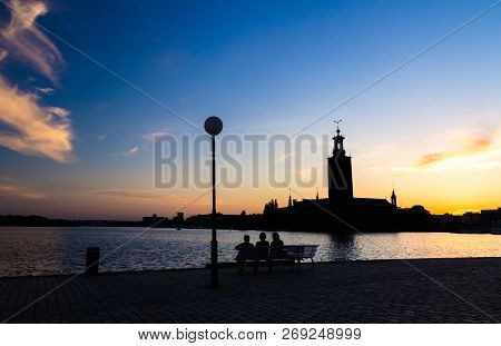 Silhouette Of Women Sitting On Bench At Promenade Quay Bank Of Lake Malaren Looking At Stockholm Cit
