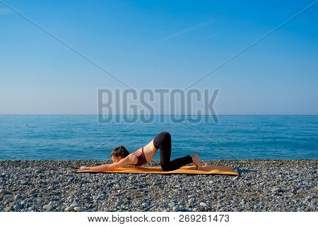 Young Slim Woman In Tight Sportswear Sitting On Orange Yoga Mat And Practicing Outdoors At Pebble Be