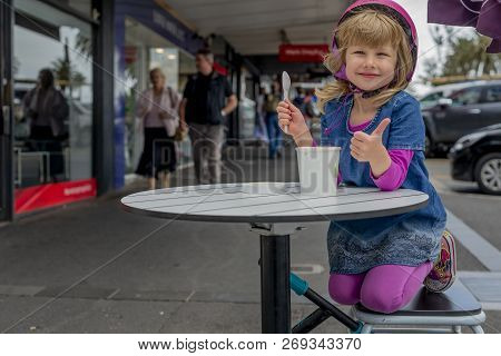 Young Girl In Pink Outfit And Pink Helmet Riding A Kick-scooter Sitting At The Outside Cafe Table Wi
