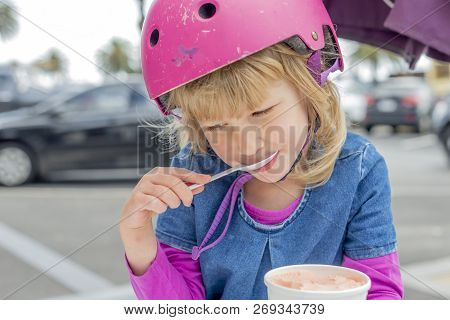 Young Girl In Pink Outfit And Pink Helmet Riding A Kick-scooter Sitting At The Outside Cafe Table Wi
